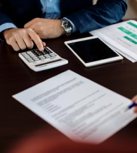 A person using a calculator on a table with documents and a tablet nearby. Another person is seen holding a pen, possibly signing a document.