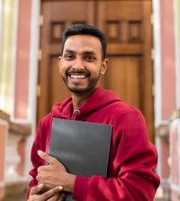 Smiling man in a red hoodie holding a black folder, standing in front of a building entrance