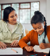Smiling woman helping a young girl writing at a table, with pencils and papers around them.